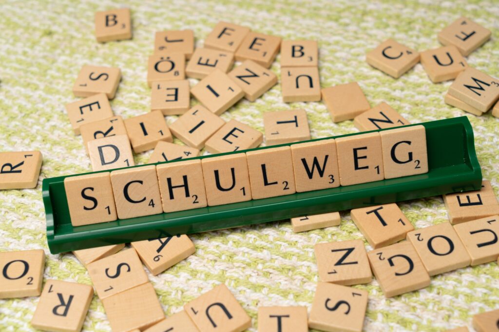 Scrabble tiles spelling 'Schulweg' on a rack, surrounded by scattered letters on a knitted background.